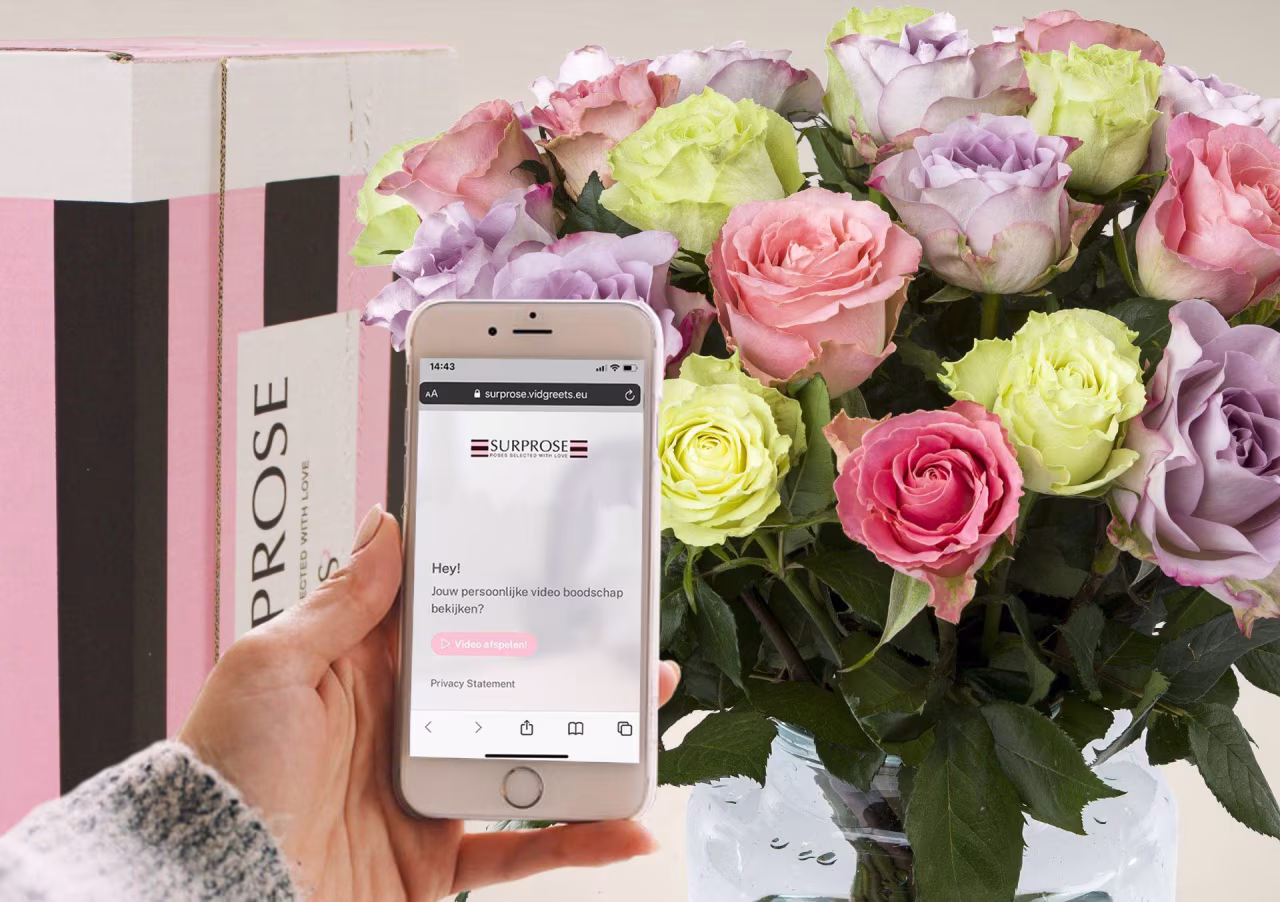 Hand houdt smartphone met website SURPROSE en tekst 'Hey! Kun je een mooie video boodschap achterlaten?'. Bos kleurrijke rozen in vaas. Primeur Surprose videoboodschap bij online bestelling rozen