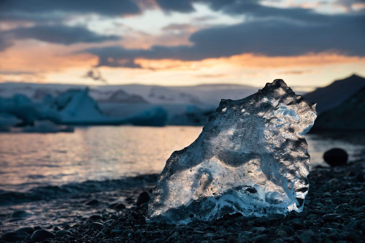 Stuk ijs op kiezelachtige oever bij meer met ondergaande zon en ijsbergen prachtige natuur en klimaatverandering