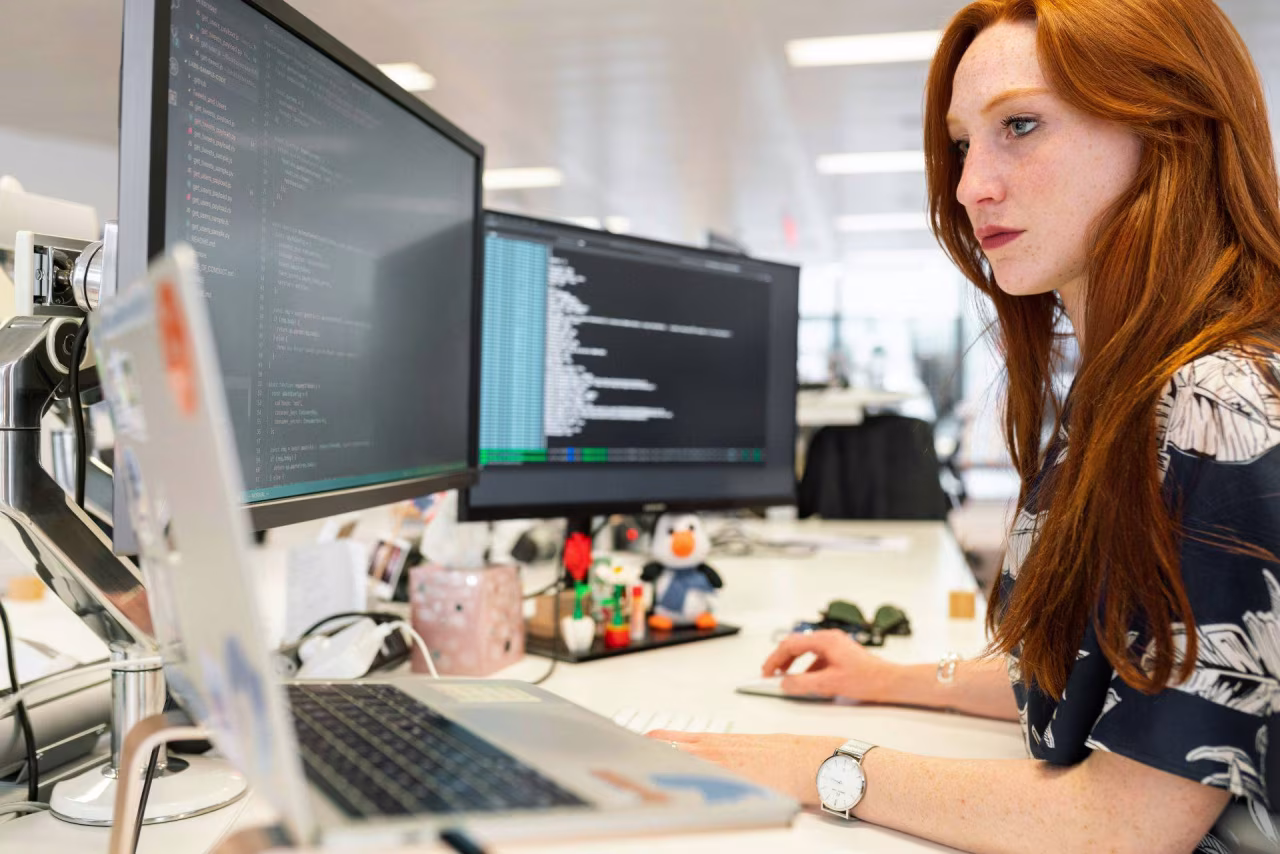Binnenkant kantoor met persoon geconcentreerd werkend aan bureau met twee computerschermen vol code en stickers op laptop, thema Vrouwen werken steeds vaker 35 uur of meer