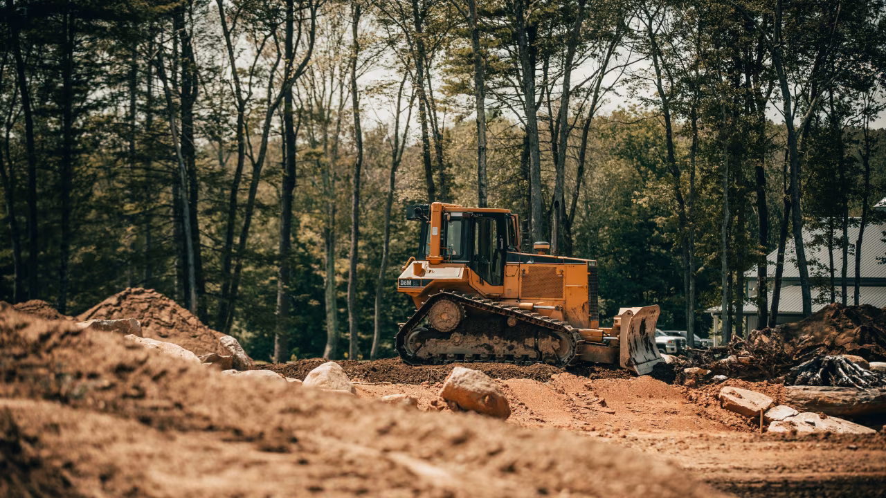 Bouwplaats in bosrijke omgeving met gele bulldozer op losse aarde, bouwmaterialen, en bomen op achtergrond Over graafmachines, gehandicapten en corona