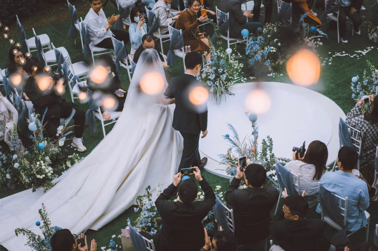 Buiten bruiloft ceremonie met bruid in lange jurk hand in hand lopend op bloemrijk pad omringd door gasten, warm licht