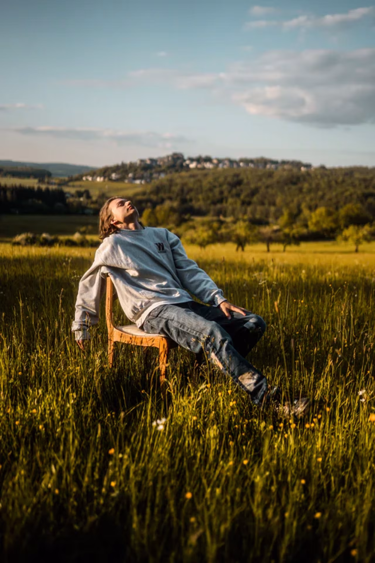 Vakantietijd, komkommertijd: persoon ontspant op houten stoel in veld met hoog gras en wilde bloemen, omgeving rust en natuur, beboste heuvel met nederzetting