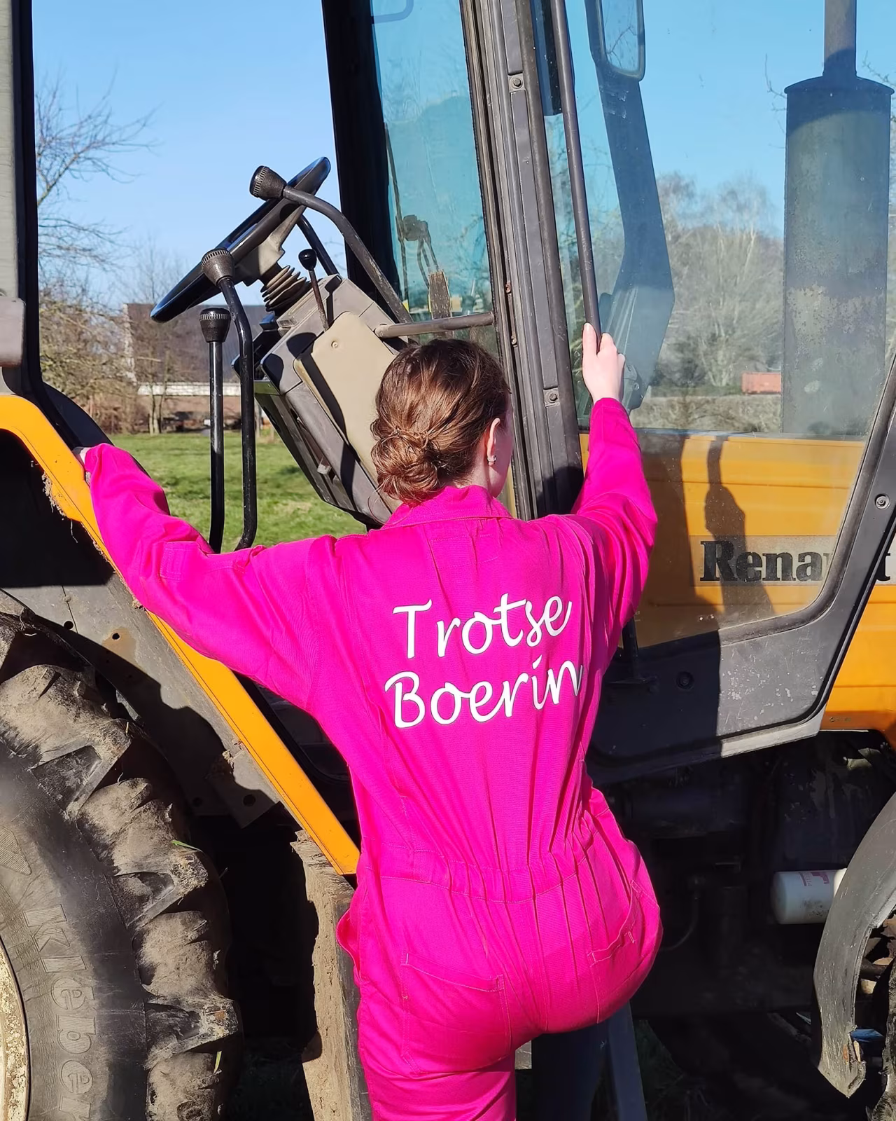 Persoon in felroze overall met tekst TROTS BOERIN kijkt naar gele tractor op groene grasvlakte onder blauwe lucht - Trekt Caroline niet een te grote overall aan