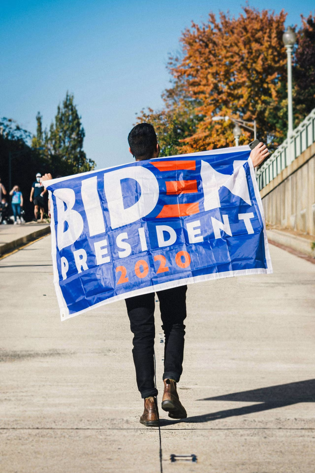 Persoon loopt over straat met blauwe vlag met grote tekst BIDEN PRESIDENT 2020 en rode strepen, politieke steun voor Joe Biden h2 titel Nooit eerder trok zittende president zich zo kort voor verkiezingen terug