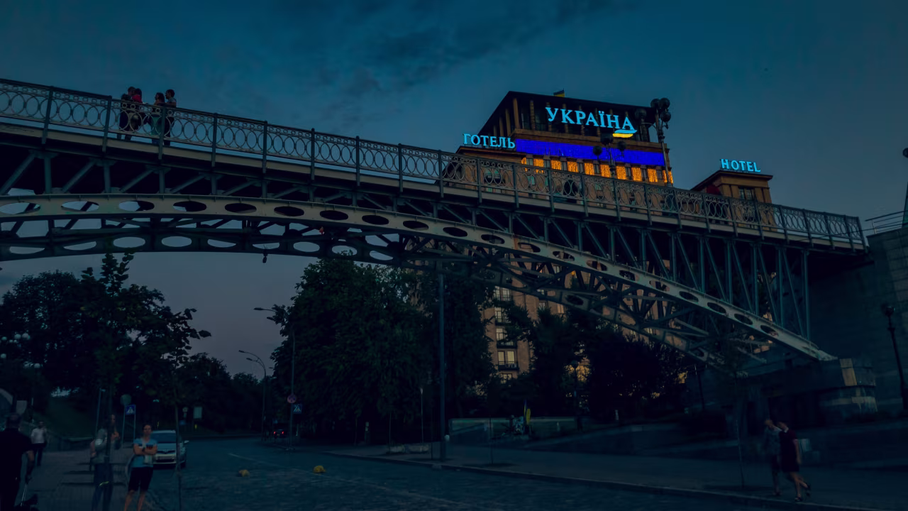 Avondfoto met metalen loopbrug en hotel met neonverlichting tekst Oekraïne in het cyrillisch, h2 titel De oorlog in Oekraïne