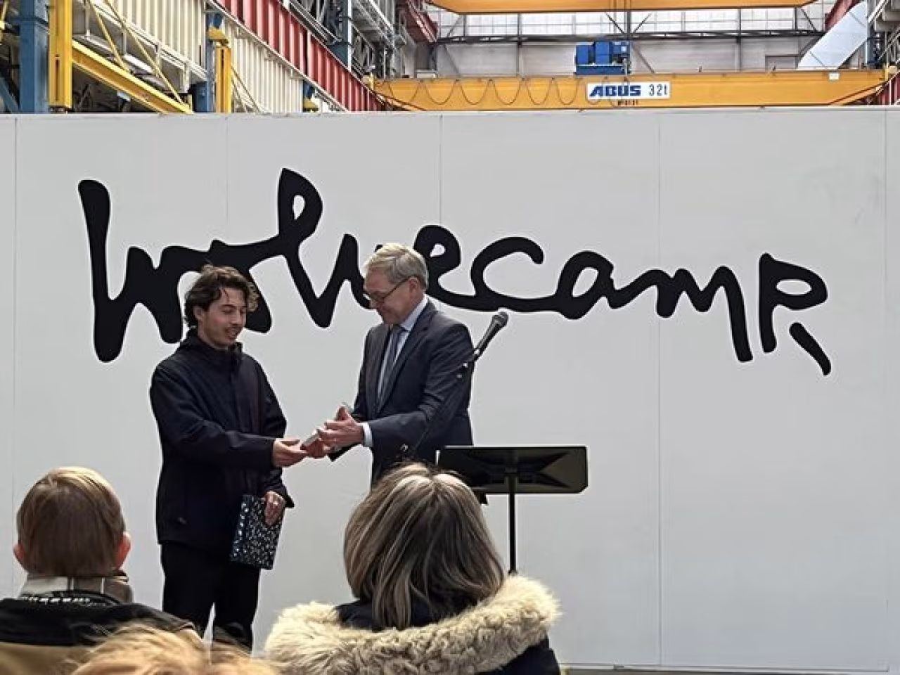 Nazif lopulissa ontvangt wolvecampprijs 2022 in fabriekshal met gele abus kraan en woord wethecamp op wand