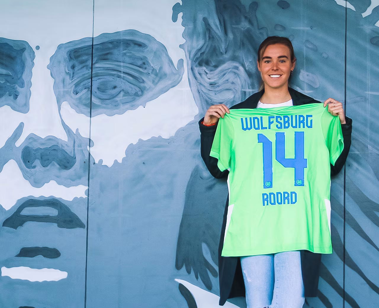 Jill Roord houdt felgroen Wolfsburg voetbalshirt nummer 14 vast voor muurschildering gezicht in context Jill Roord heeft blik alweergericht op VfL Wolfsburg