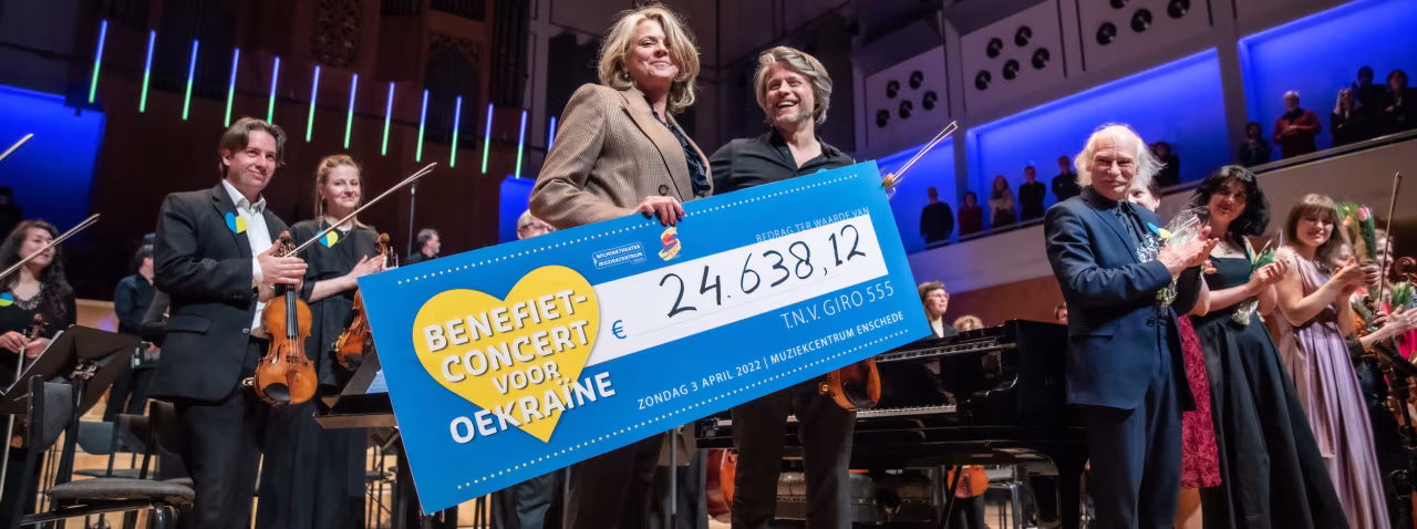 Benefietconcert voor Oekraïne toont cheque 24.638,12 euro op podium in muziekcentrum met muzikanten op achtergrond