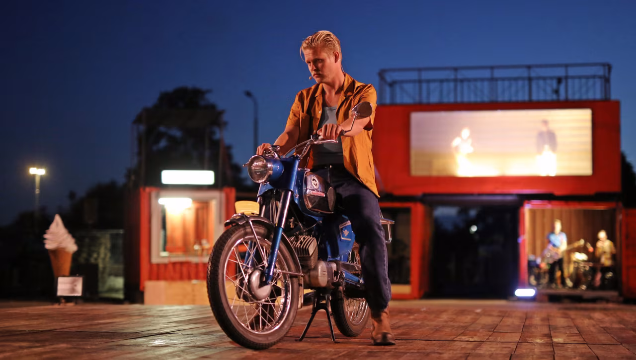 Scène bij avondlicht van motor met ruiter voor rood-oranje gebouw tijdens openluchtvoorstelling 'Spaghettirellen' cultuurclash Tukkers Italianen