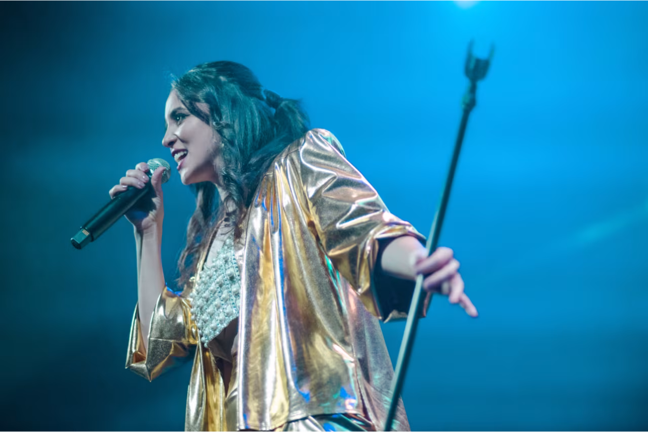 Karsu live op podium tijdens gelijkspel concert met microfoon in linkerhand en metallic jas blauwe podiumverlichting