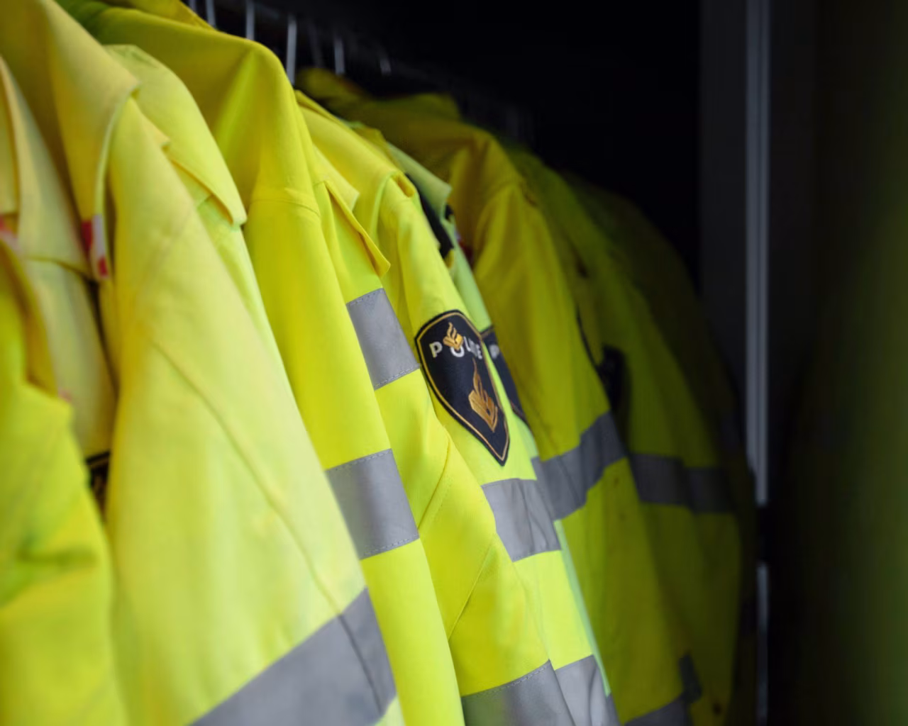 Rij geelgroene politie veiligheidsjassen met reflecterende strepen in donkere kleedruimte inrekenvraag politie en veiligheid