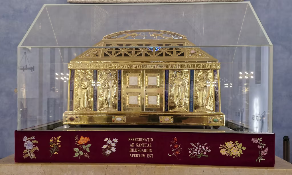 Gouden reliekhouder met gedetailleerde reliëfs achter glas met tekst PEREGRINATIO AD SANCTAE HILDEGARDIS APERTUM EST beentjes