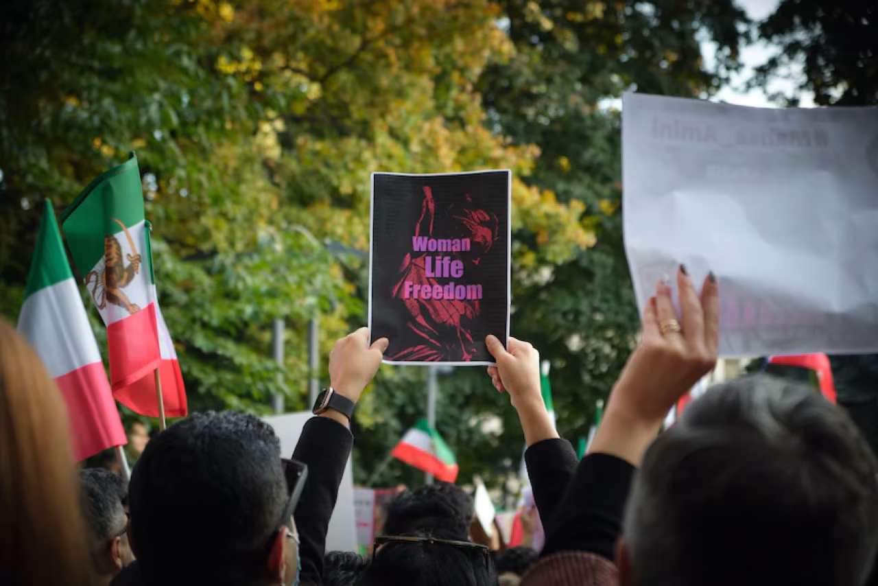 Protestmenigte buiten met borden, centraal ‘Woman Life Freedom’ bord in roze, Iraanse vlaggen zichtbaar, bericht over Iraanse Moraalpolitie