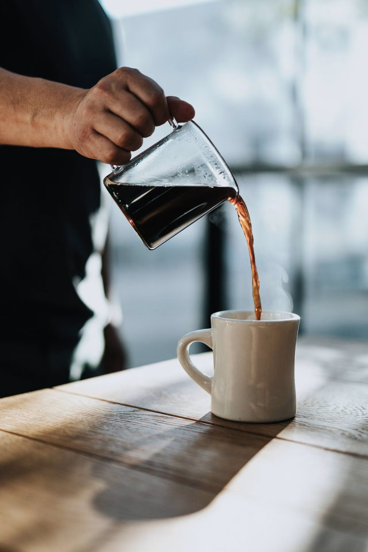 Hand schenkt dampende koffie in witte mok op houten tafel sfeer ochtendritueel leut TKKR